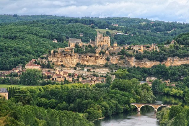 Beynac Castle in Dordogne Beynac Castle in Dordogne surrounded by green landscape with the Dordogne bridge flowing through it. There's a bridge over the river.