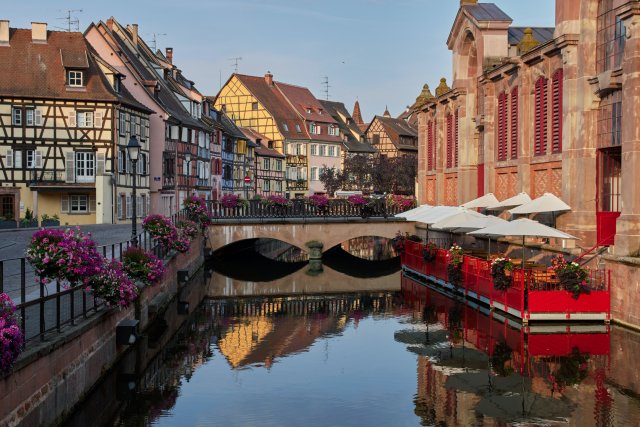 The canal running through Little Venice in Colmar, Alsace. There are colorful half-timbered houses to the left of the canal. The sky is blue.