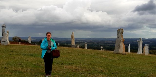 Trip Planner Emilie in La Vallée des Saints in Brittany France Just For You trip planner Emilie in the Saints' Valley in Brittany. There are large sculptures of saints on the grassy field behind her. It's a cloudy day. Emilie is smiling.
