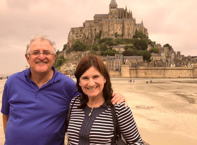 Travelers Lucelle and Alan from South Africa in front of Mont Saint Michel A smiling man and woman standing in front of Mont Saint Michel Islet in Normandy