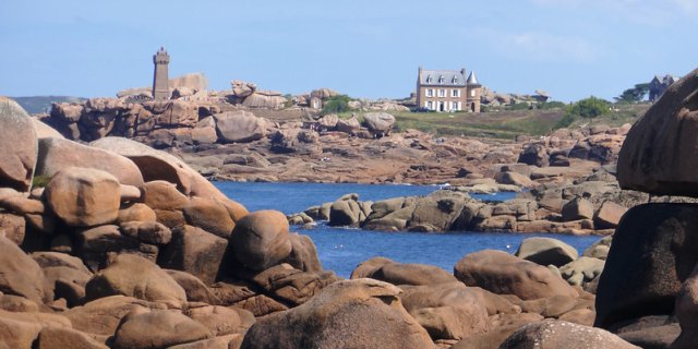 The pink boulders on the Pink Granite Coast in Brittany, with a house and lighthouse in the distance, beyond the sea.