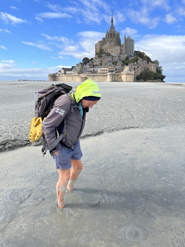 A tour guide with bare feet paddling in the shallow water of the bay of Mont Saint Michel, with Mont Saint Michel islet and abbey in the background. The sky is blue with a few clouds.