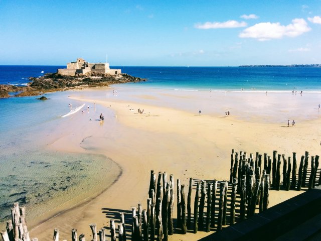 The beautiful beach of Saint-Malo in Brittany. The sand is golden yellow and stretches out to the fortress in the turquoise sea. The weather is sunny.