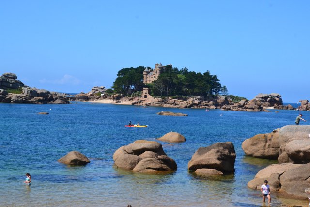Saint-Guirec beach in Brittany. There are big boulders in the sea, and people paddling, with an island with a building on it in the distance.