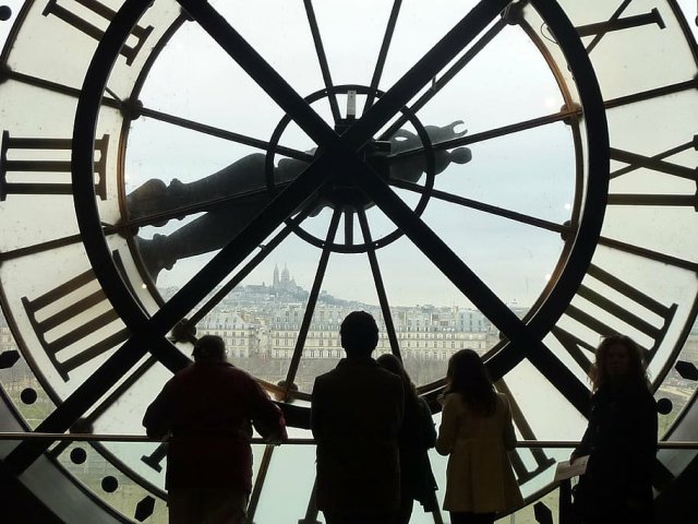 The big glass clock inside the Musee D'Orsay in Paris. There are people standing in front of it looking through the glass.