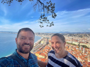 A smiling couple on their honeymoon in the south of France. Nice and the Mediterranean sea are in the background. The sky is blue and it's a sunny day.